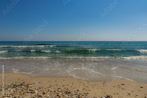 The surf of the blue turquoise sea with white waves on the sandy beach against a bright clear sky.