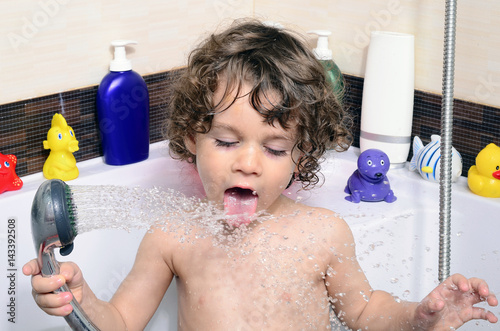 Beautiful toddler taking a bath in a bathtub with bubbles. Cute kid washing his hair with shampoo in the shower and splashing water everywhere. Boy playing with toys in the tub photo