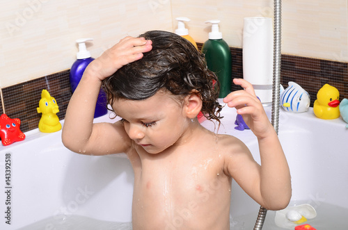 Beautiful toddler taking a bath in a bathtub with bubbles. Cute kid washing his hair with shampoo in the shower and splashing water everywhere. Boy playing with toys in the tub photo