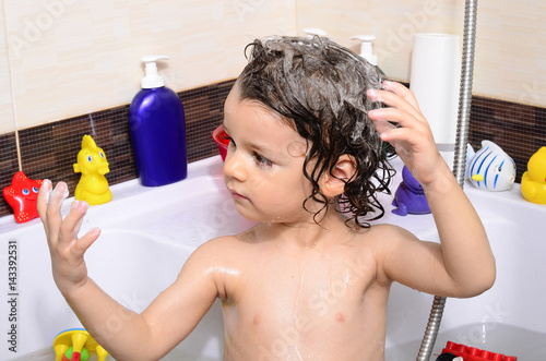 Beautiful toddler taking a bath in a bathtub with bubbles. Cute kid washing his hair with shampoo in the shower and splashing water everywhere. Boy playing with toys in the tub photo