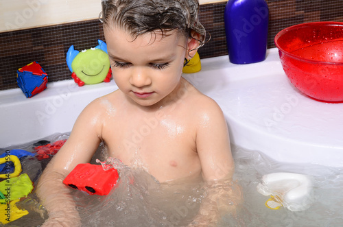 Beautiful toddler taking a bath in a bathtub with bubbles. Cute kid washing his hair with shampoo in the shower and splashing water everywhere. Boy playing with toys in the tub photo