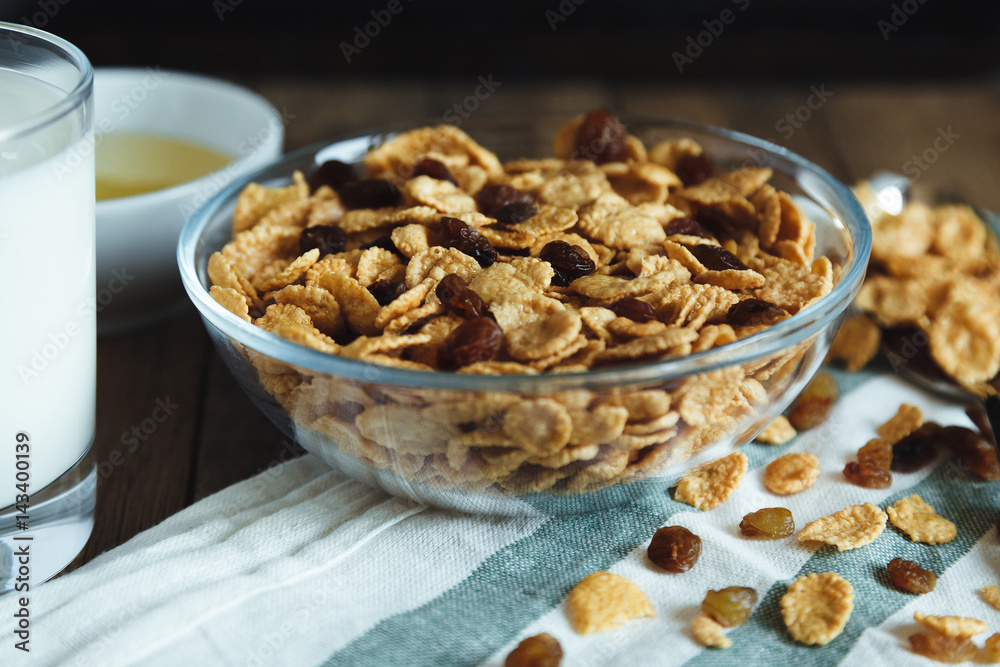 Cornflakes with raisins in bowl, honey and glass of milk on the table with towel Cornflakes with raisins in bowl, honey and glass of milk on the table with towel
