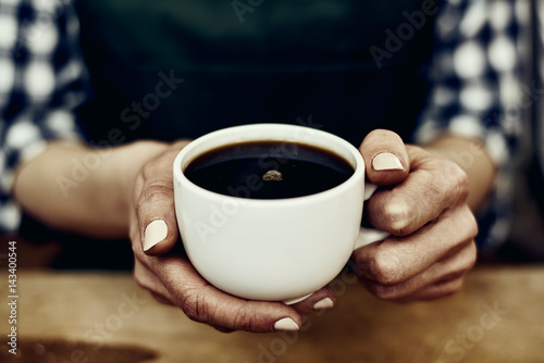 Female waitress in apron holding hot coffee mug indoors.