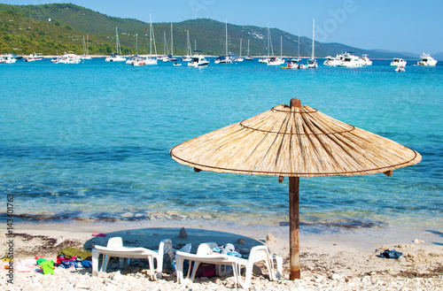 One yellow straw umbrella and two chairs on the pebble sandy beach. Many yachts in the azure blue sea lagoon in a bay near the coast of an island. Blue cloudless sky. Dugi otok, Croatia
