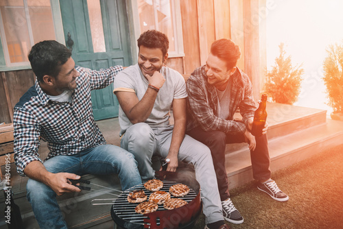 Three young men drinking beer and smiling while sitting on porch and making barbecue