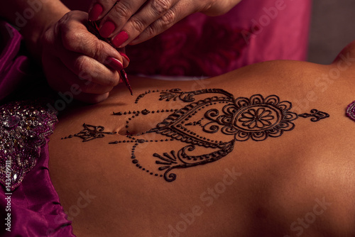 White woman a mehendi artist paints an ornament of henna on an eastern beautiful girl’s stomach photo