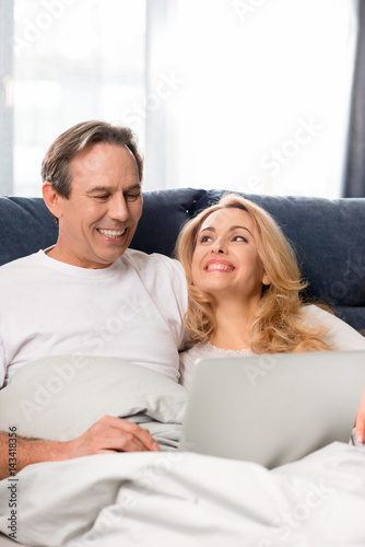 Happy middle aged couple using laptop and lying on bed at home