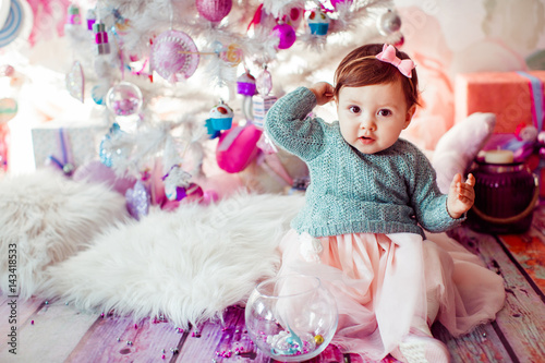 Pretty little girl in mint dress sits on fluffy carpet before Christmas tree