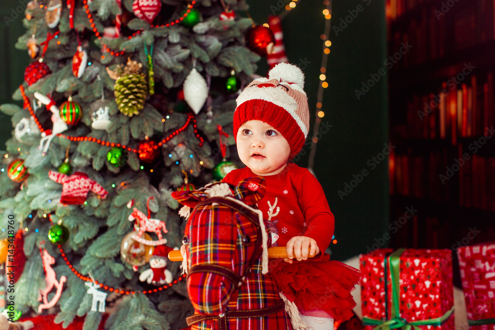 Obraz premium Little girl in woolen hat sits on toy horse before Christmas tree