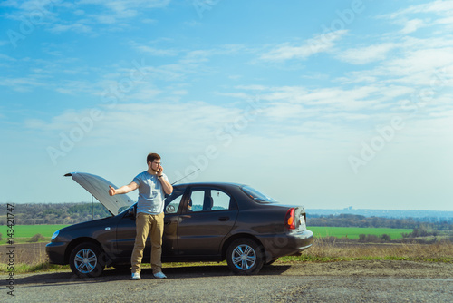 Angry young man waiting a help while sitting near the broken car