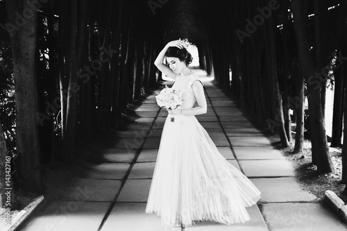 Bride in light summer dress holds her hand up posing on path in park