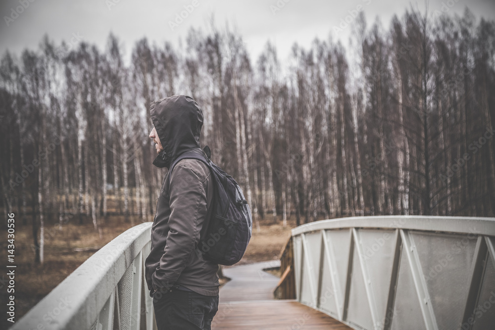 Fototapeta premium Sad, lonely young man on the footbridge in the cold, rainy spring day. Gloomy atmosphere.
