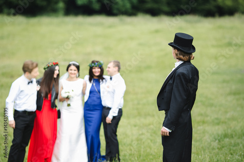 Bride and friends stand on the lawn befoe groom in black tailcoat and topper