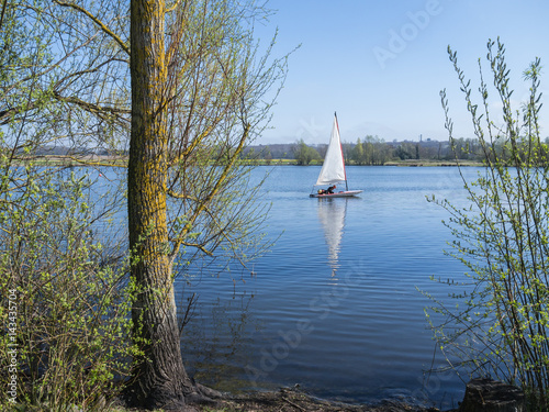 A sailing dinghy and its reflection on a peaceful blue lake, conningbrook lakes country park, with trees in the foreground.