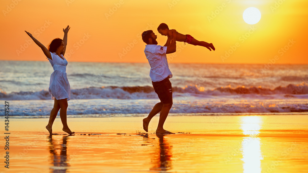 Fototapeta premium Happy family - father, mother, baby son walk with fun along edge of sunset sea surf on black sand beach. Active parents and people outdoor activity on summer vacations with children on Bali island