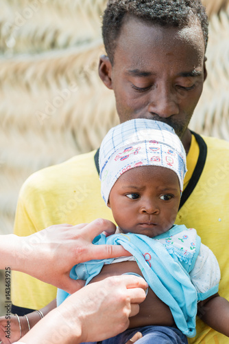 Female Caucasian doctor listening breath and heart beat of little African baby with stethoscope.Father holding the baby, unrecognizable female doctor