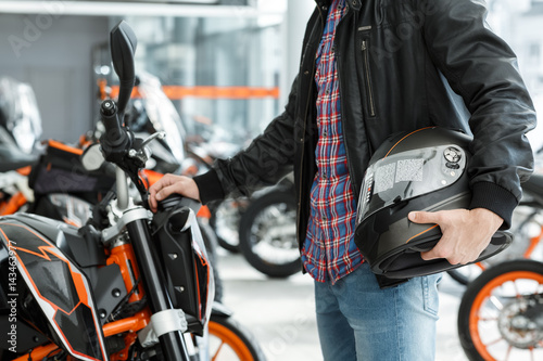 Important detail. Cropped shot of a man wearing leather jacket standing near his newly bought motorbike holding a black helmet at the motorcycle salon