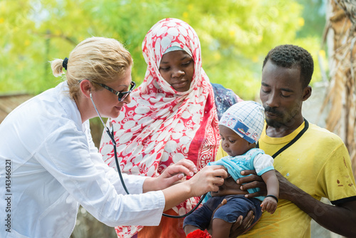 Female Caucasian doctor listening breath and heart beat of little African baby with stethoscope.Father holding the baby, mother looking at baby photo