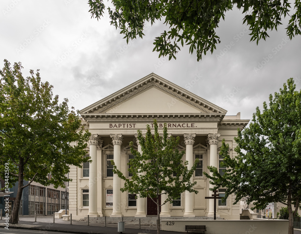 Auckland, New Zealand - March 1, 2017: Greek-Roman architecture for beige facade with triangular frieze of Baptist Tabernacle in Queen Street. Green vegetation and line of pillars.