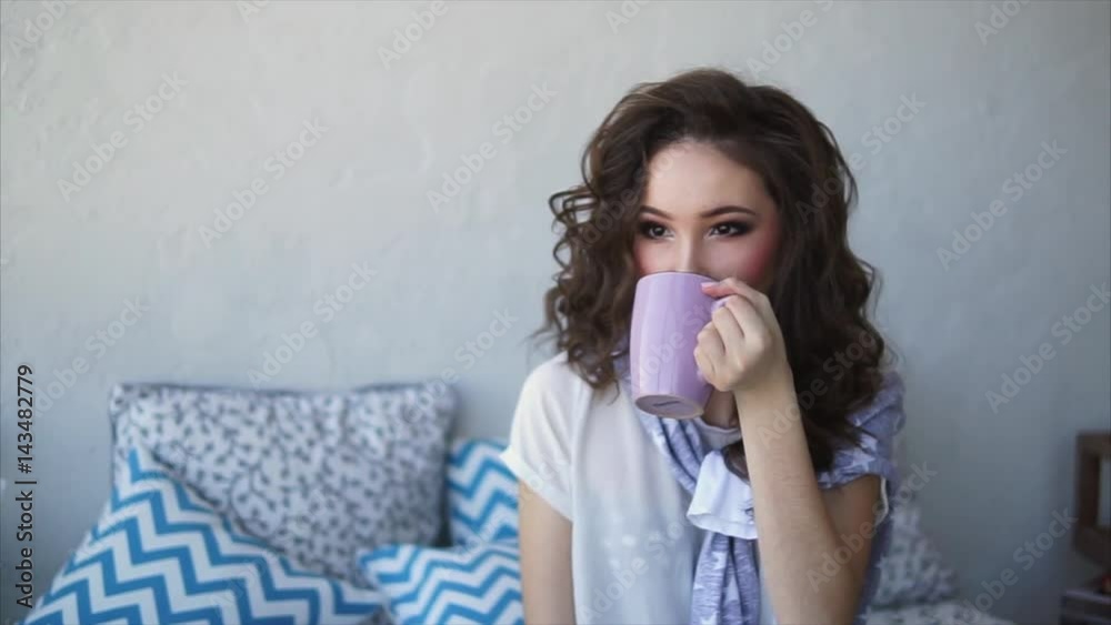An adult happy woman, dressed in pajamas, drinks morning coffee in the bedroom. A girl with curly brown hair enjoys breakfast in bed on which lie pillows. An adult happy woman, dressed in pajamas, drinks morning coffee in the bedroom. A girl with curly brown hair enjoys breakfast in bed on which lie pillows.