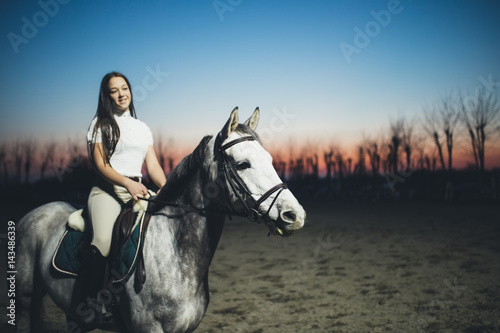 Beautiful young teenage girl riding a white horse at sunset. Selective focus on horse's head.