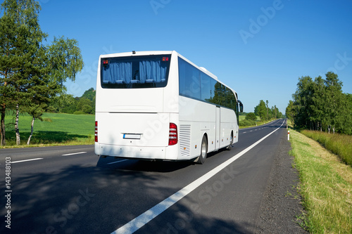 The white bus moving on asphalt road lined avenue of trees in a rural landscape on a bright sunny day