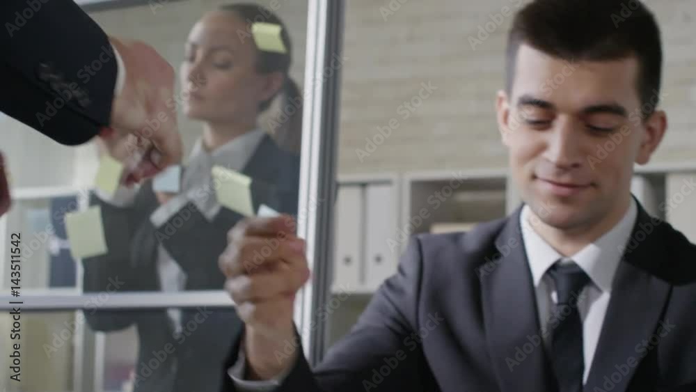 Handsome office worker sitting at desk in office and greeting colleague with handshake while business woman putting sticky notes on glass wall Handsome office worker sitting at desk in office and greeting colleague with handshake while business woman putting sticky notes on glass wall