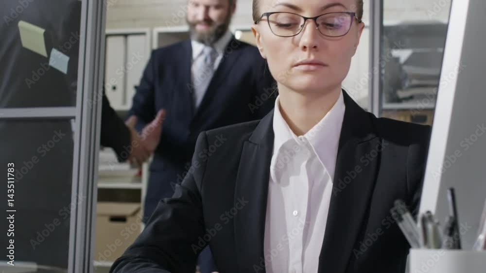 Tilt up of smart businesswoman in eyeglasses and formal suit typing on computer at desk while two business partners shaking hands in background in office Tilt up of smart businesswoman in eyeglasses and formal suit typing on computer at desk while two business partners shaking hands in background in office