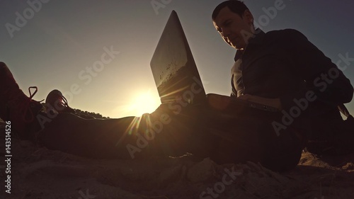 man businessman freelancer working behind laptop sitting on beach silhouette in the sun freelancing