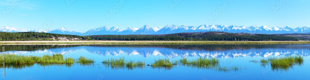 Panorama of the spring flood of the river. Snow-capped peaks of the Eastern Sayan Mountains are reflected in the blue water of the river
