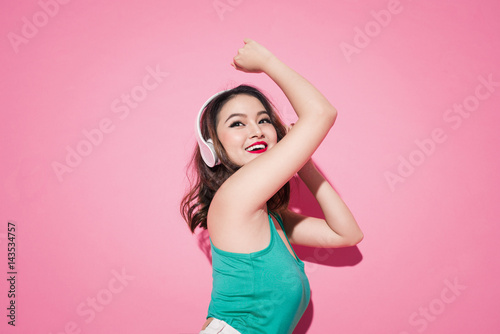 Beautiful asian girl with professional makeup and stylish hairstyle singing and dancing while listening to music on pink background.