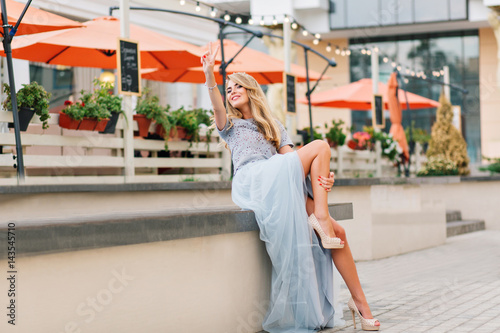 Attractive girl with long blonde hair in blue long tulle skirt having fun on terrace background. She keeps hand on leg and smiling to camera © Look!