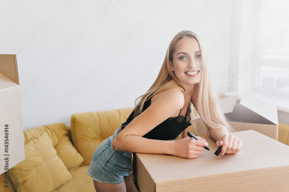 Portrait of gorgeous girl going to write label on box, packing, relocating to new home, flat, apartment, house. Happy blonde holding marker in hand in room with yellow sofa. Wearing black top, shorts. Portrait of gorgeous girl going to write label on box, packing, relocating to new home, flat, apartment, house. Happy blonde holding marker in hand in room with yellow sofa. Wearing black top, shorts.