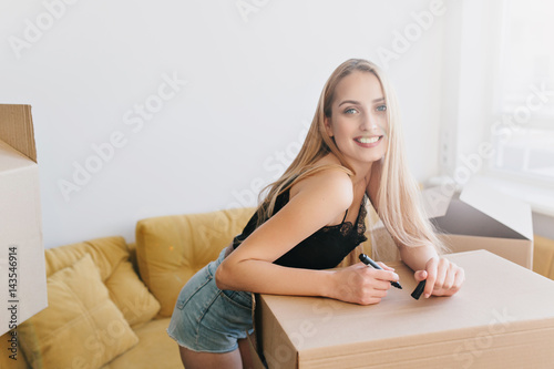 Portrait of gorgeous girl going to write label on box, packing, relocating to new home, flat, apartment, house. Happy blonde holding marker in hand in room with yellow sofa. Wearing black top, shorts.