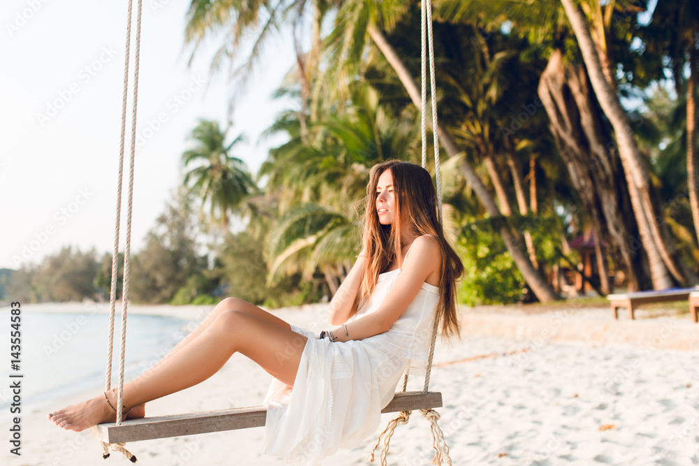 Sensual tender girl sitting on a swing wearing white dress. Girl has her eyes closed. She has long dark hair. She has bracelets on her arm and leg. The swing is on the beach with green palms.