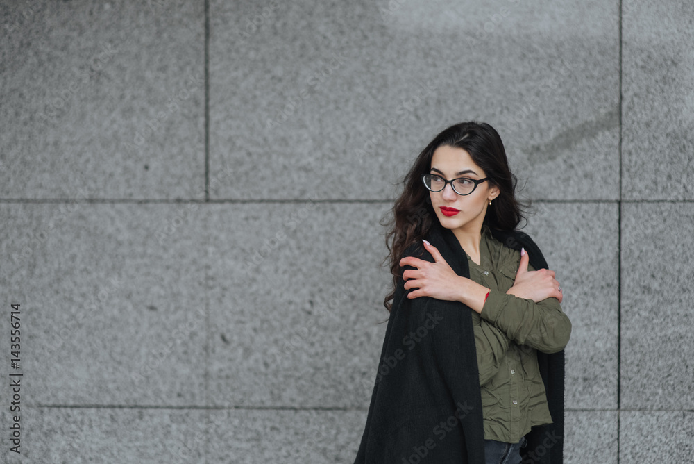 Fashion consept: beatiful young girl with long hair, glasses, red lips standing near modern wall wearing in green suit and grey jeans. She wanted talking on phone close eyes