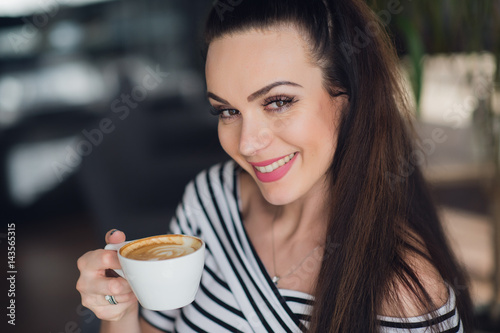 A close-up portrait of a woman with perfect smile holding a cup of cappuccino or coffee. White teeth and makeup.