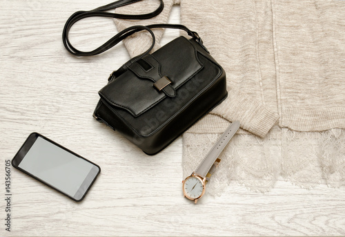 Black handbag, watch and mobile phone on a beige cardigan. Fashionable concept, top view