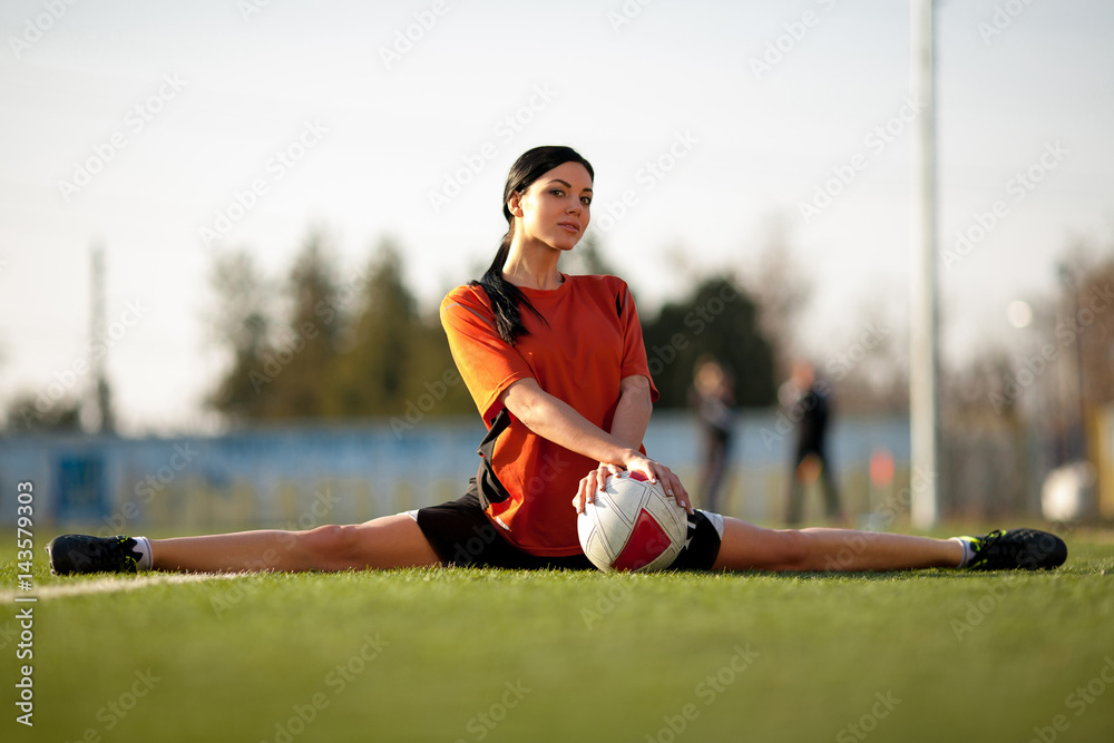 Obraz premium Girl in football uniform with ball, sitting in gymnastic twine on football field.
