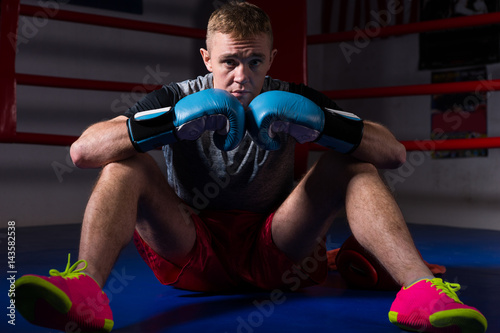 Young male boxer in boxing gloves sitting in regular boxing ring