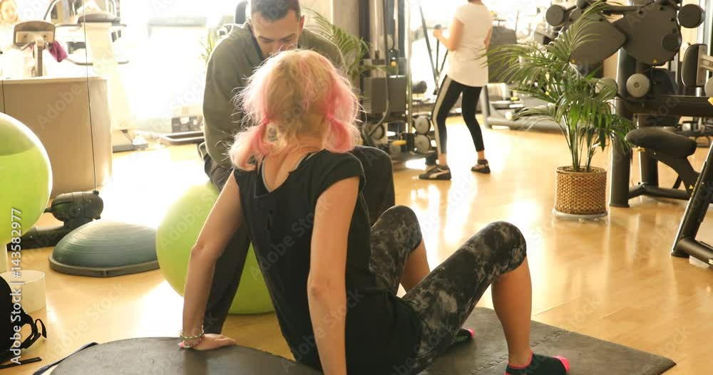 Instructor male flirting with athlete woman training her biceps, view from behind. Woman in sportswear training arms and shoulders on vibration plate machine with help from Personal trainer of gym.