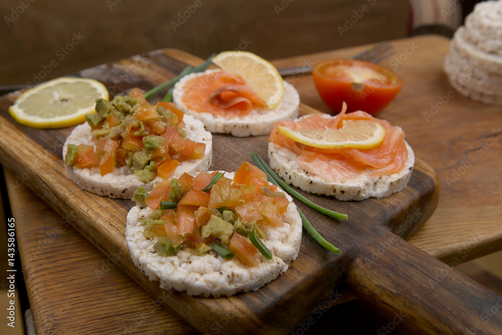 Snacks from rice cookies, salmon, tomatoes, avocado on cutting board and wooden table. Snacks from rice cookies, salmon, tomatoes, avocado on cutting board and wooden table.
