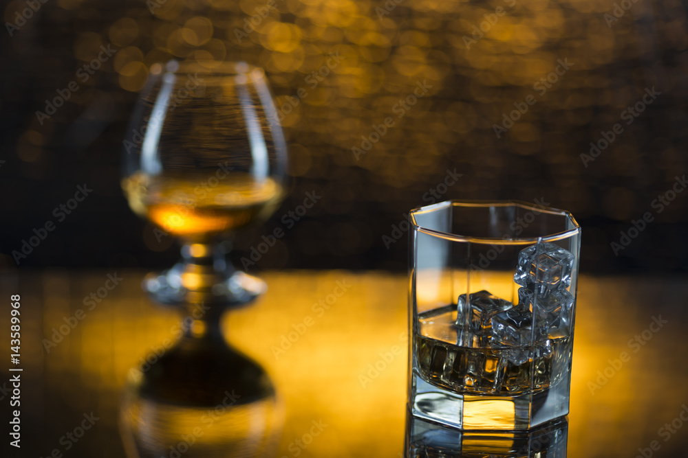 Glass o whisky with ice and wineglass of coghac on yellow bokeh background Glass o whisky with ice and wineglass of coghac on yellow bokeh background