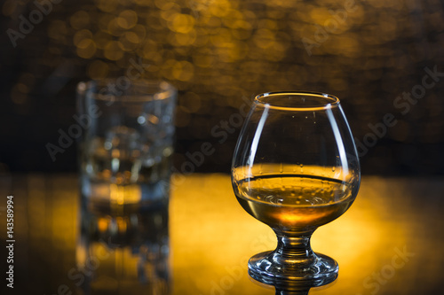 Glass o whisky with ice and wineglass of coghac on yellow bokeh background