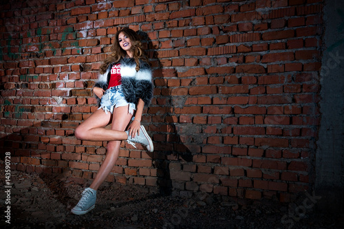 Stylish cool cool girl in short shorts with a red T-shirt and fur coat, with curly long hair posing in an abandoned building with brick walls