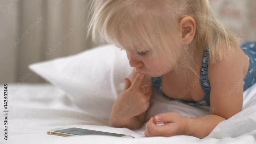 Girl lying on white bed and playing smartphone with her finger in the mouth Girl lying on white bed and playing smartphone with her finger in the mouth