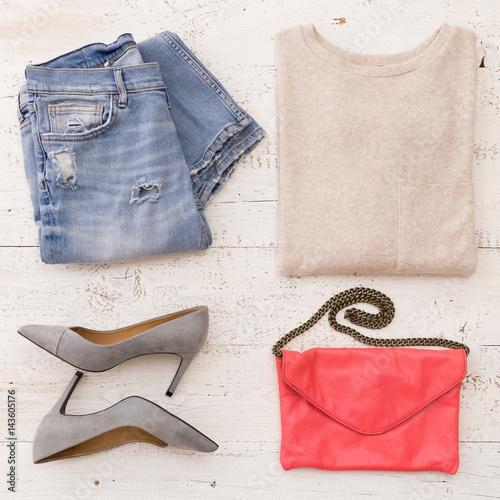 Top view on woman's fashion clothes: blue jeans, sweater, heels, pink leather clutch on white wooden background.
