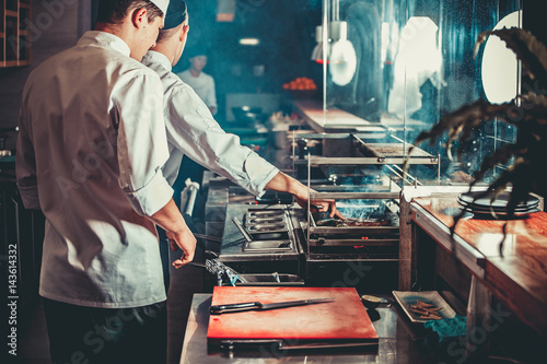 Food concept. Two chefs in white uniform monitor the degree of roasting meat with hand in interior of modern restaurant kitchen. Preparing traditional beef steak on barbecue oven from stainless steel