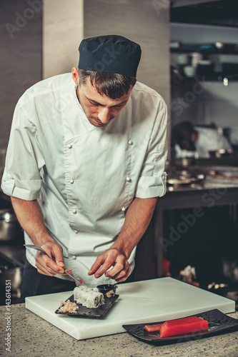 Food concept. One young white chef dressed in white uniform decorate ready dish in restaurant. He is working on maki rolls. Preparing traditional japanese sushi set in interior of modern kitchen © Goinyk