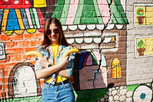 Beautiful fun teenage girl with bananas at hands, wear yellow t-shirt, jeans and sunglasses near graffiti wall.